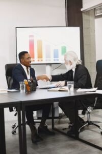 Two diverse businessmen in suits shaking hands over a desk in a modern office.